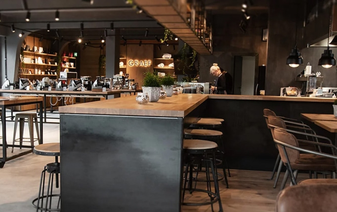 Intérieur d'un café avec un bar, des tables et des chaises, une personne et des plantes.