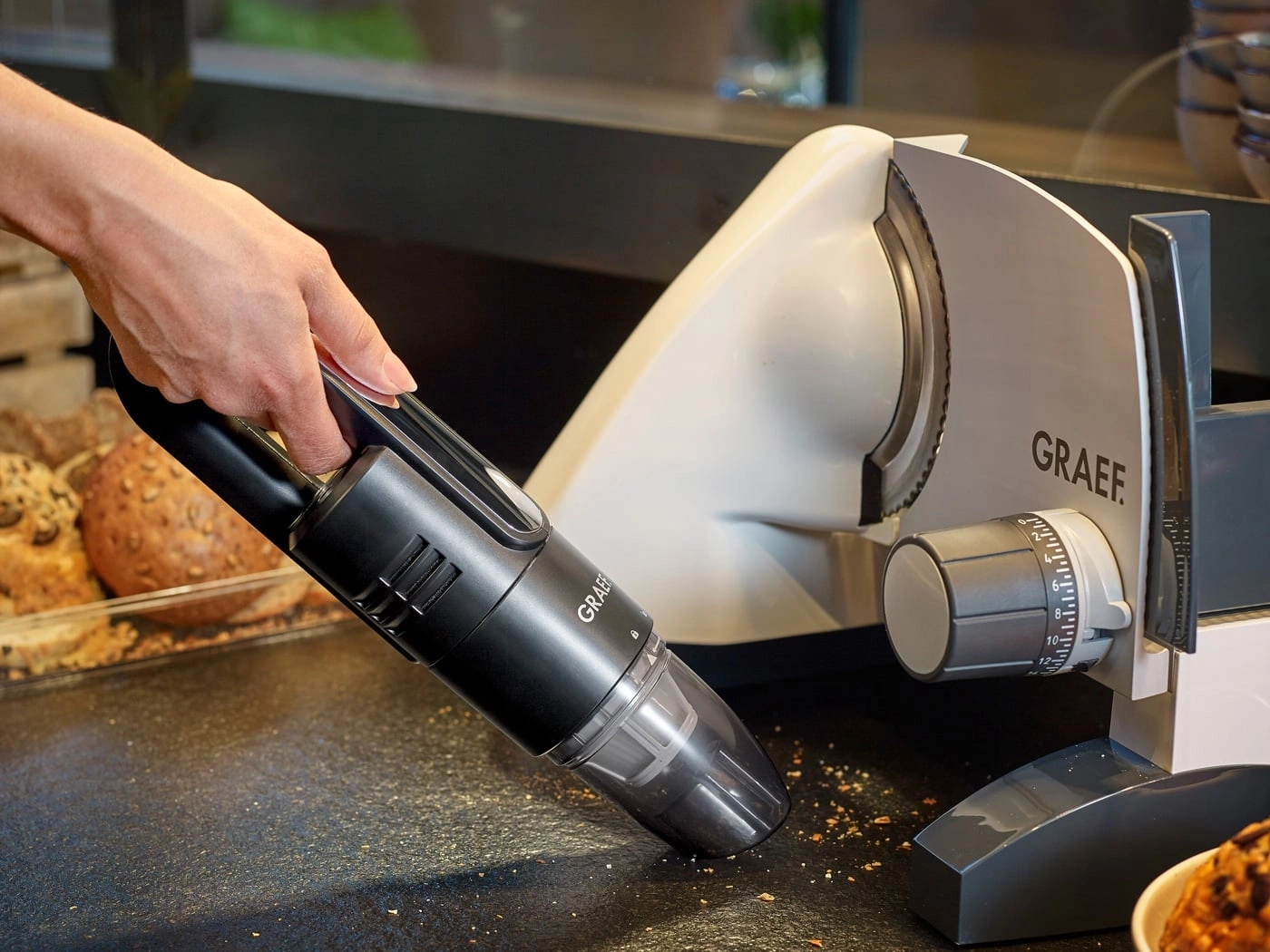 Main tenant un aspirateur au-dessus des miettes de pain à côté d'une trancheuse GRAEF.
