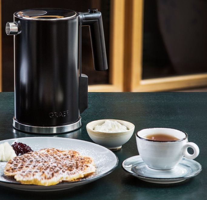 Schwarzer Wasserkocher, Waffel, Sahne, Marmelade und Tee auf dem Tisch.