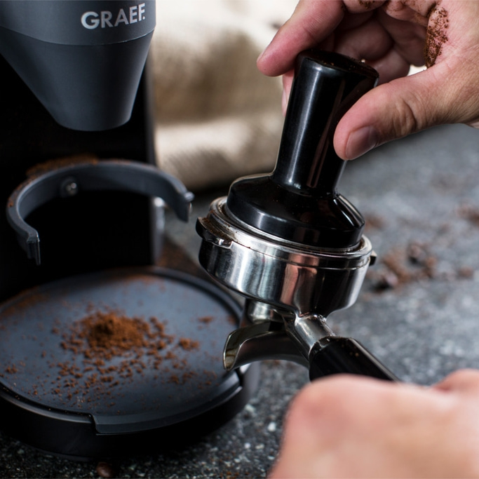 Person pressing coffee grounds with a tamper into a portafilter, close-up shot.