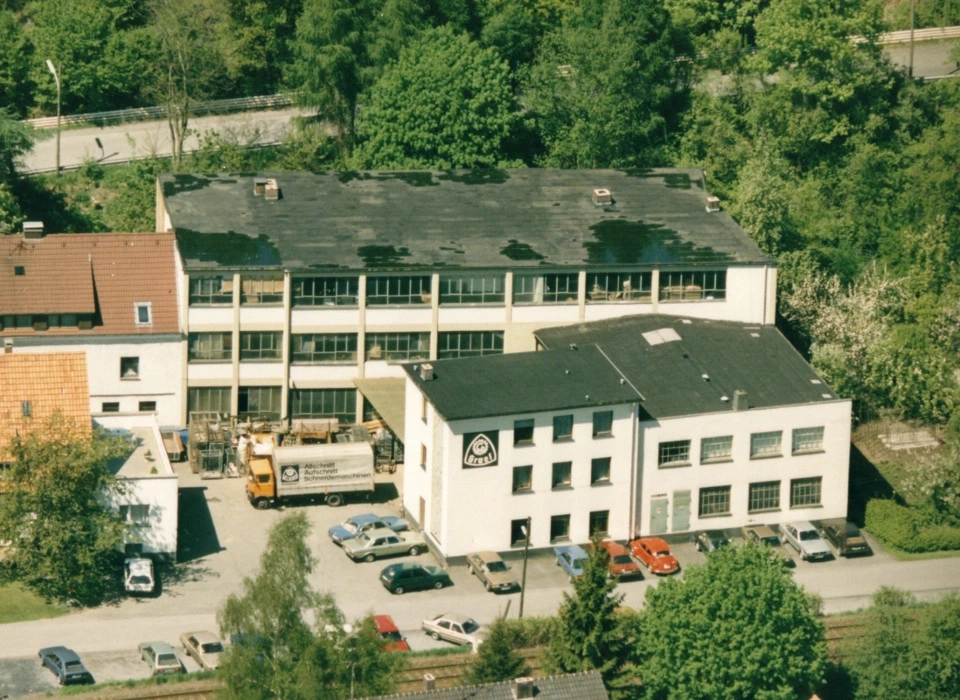 Luftaufnahme eines Fabrikgebäudes mit Parkplätzen und Bäumen in der Nähe.