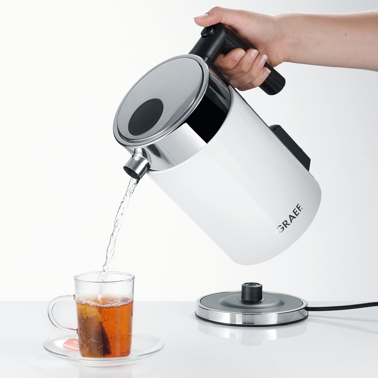 Eine Hand gießt Wasser aus einem weißen Wasserkocher in eine Teetasse.