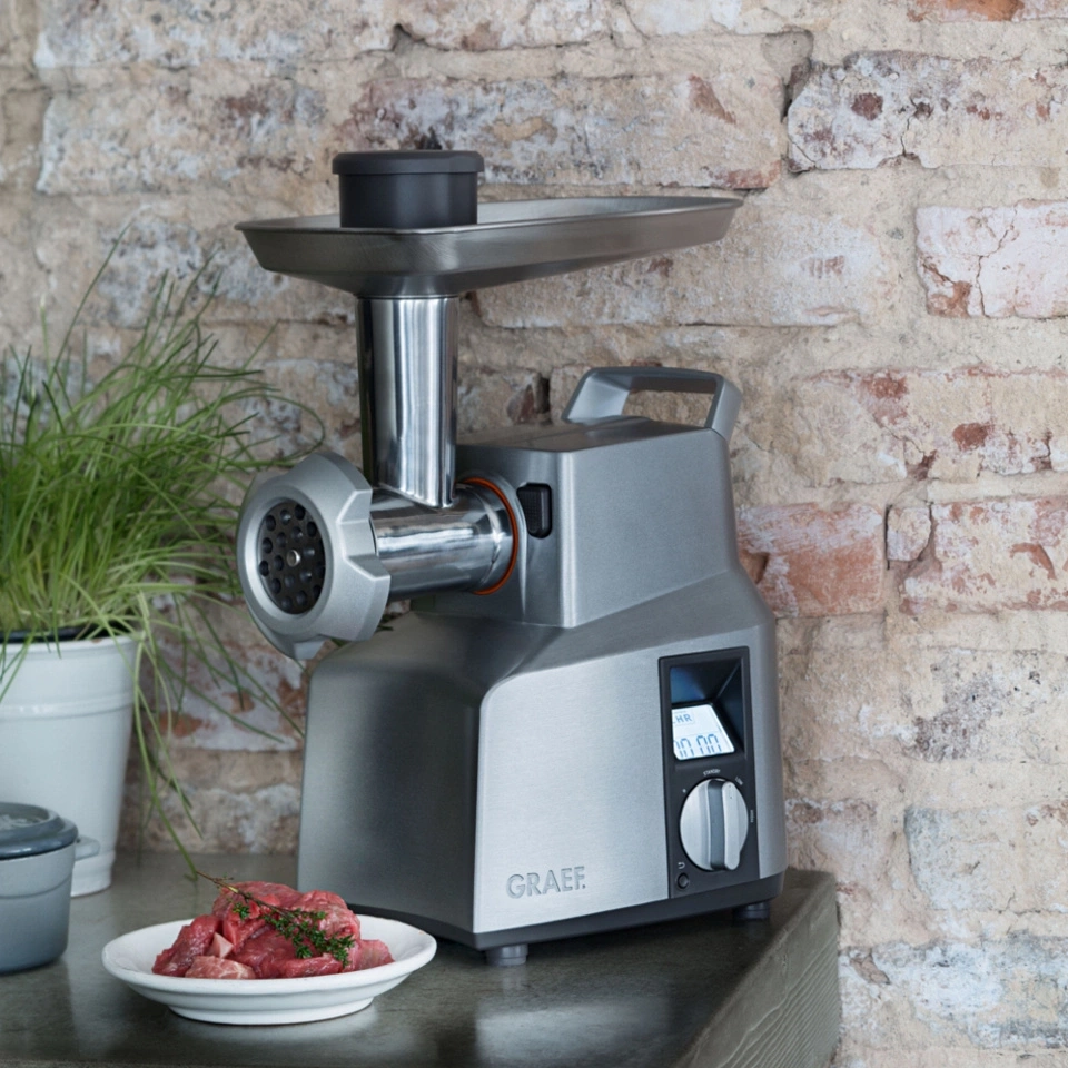 Meat grinder with meat on a plate in front of a brick wall background.