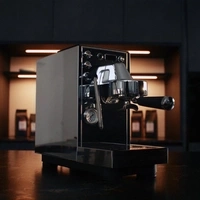 Eine glänzende Espressomaschine steht auf einem dunklen Tisch, im Hintergrund sind Kaffeepackungen.