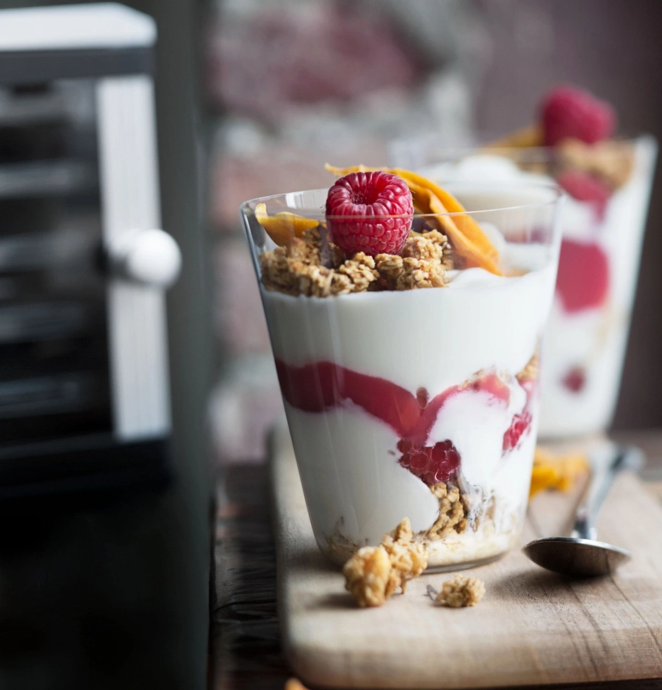 Joghurt-Dessert mit Himbeeren, Granola und Mango-Chips in Gläsern auf Holzbrett