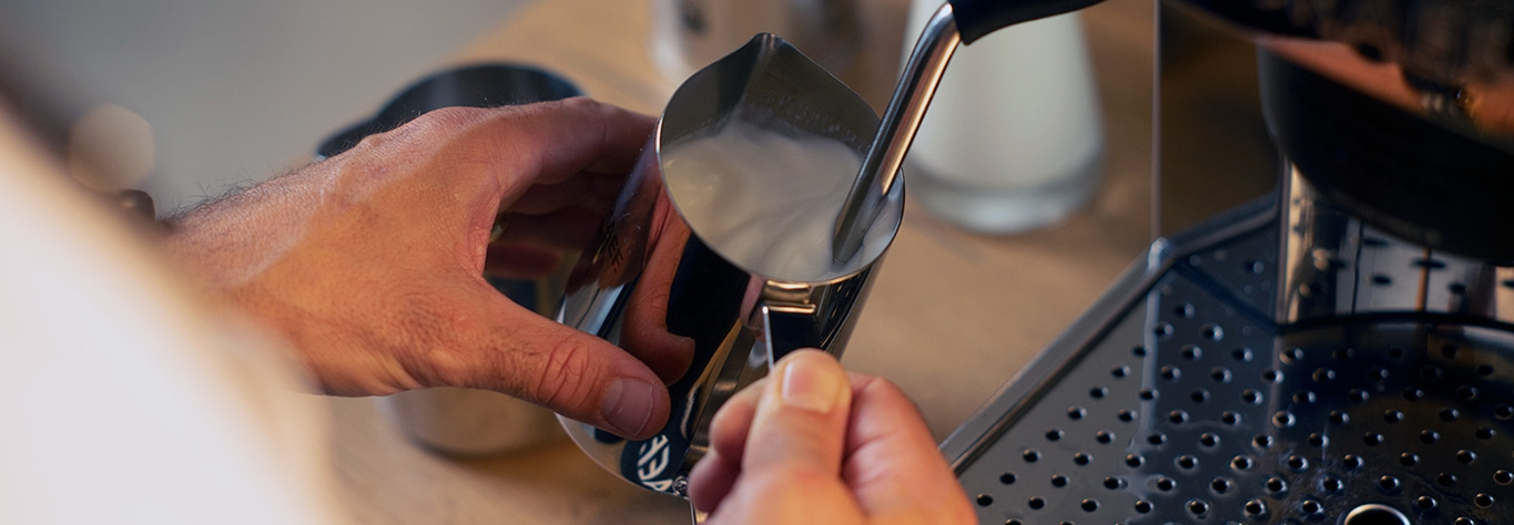Nahaufnahme einer Person, die Milch in einem Metallkrug aufschäumt, um Kaffee zuzubereiten.
