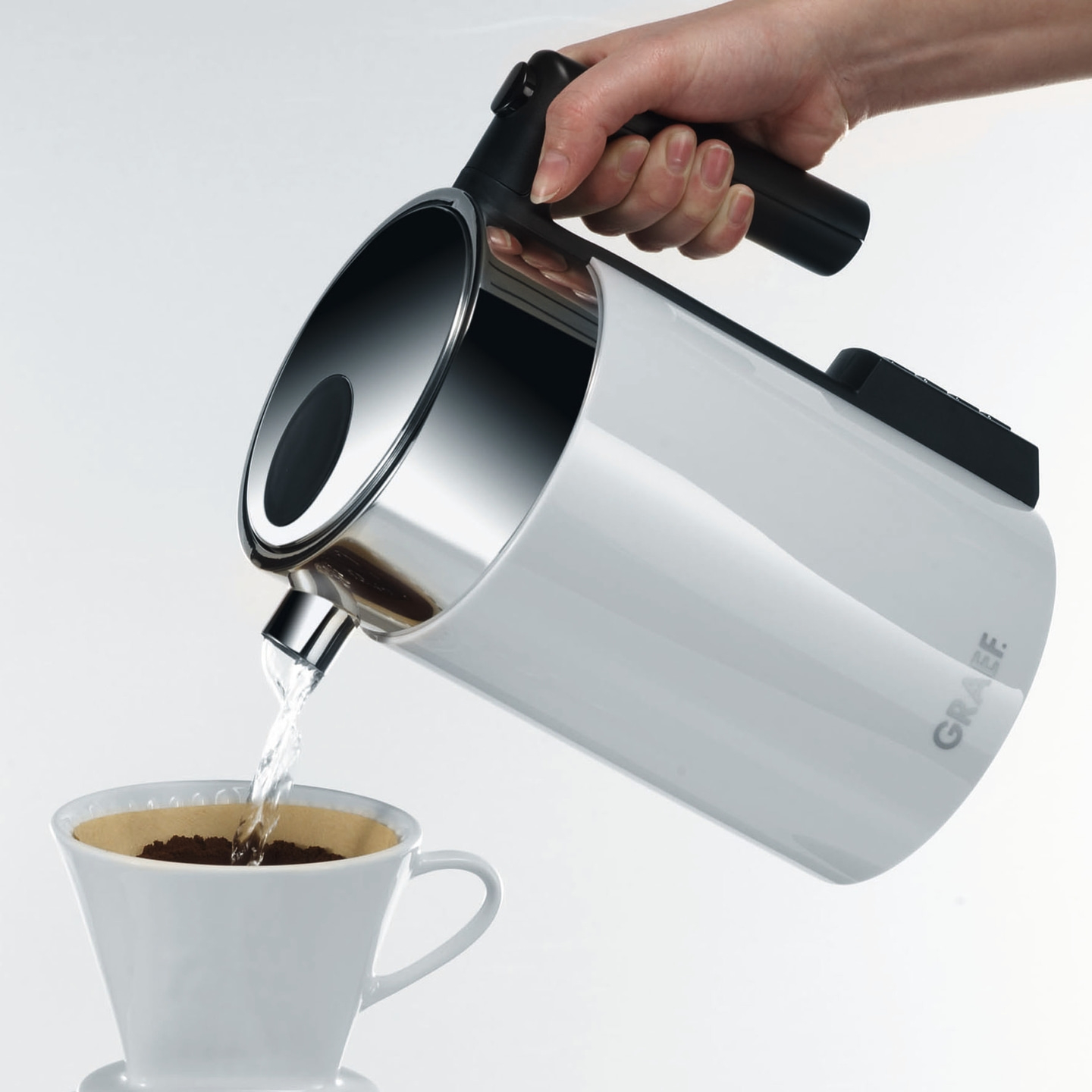 Kaffeekanne gießt Wasser in eine Tasse mit Kaffeepulver, Hand hält Kanne.