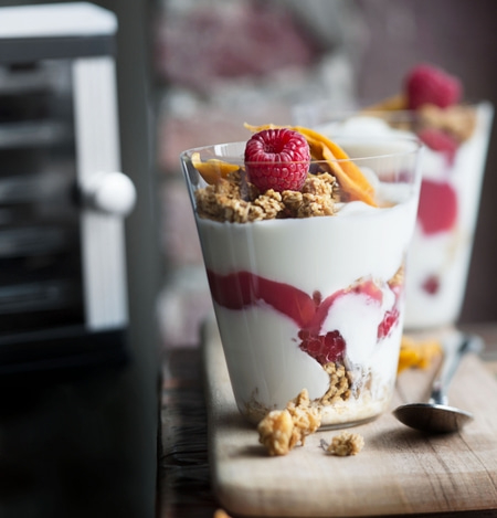 Joghurt-Dessert mit Himbeeren, Granola und Mango-Chips in Gläsern auf Holzbrett