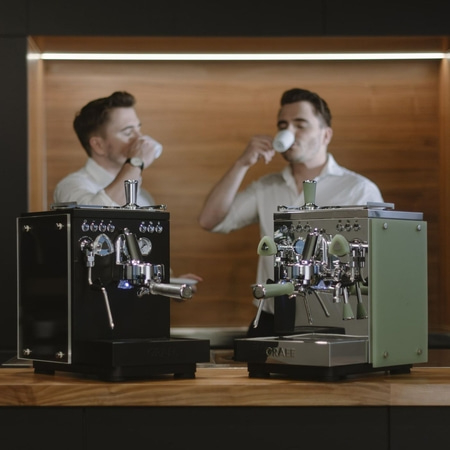 Two men drinking coffee next to espresso machines.