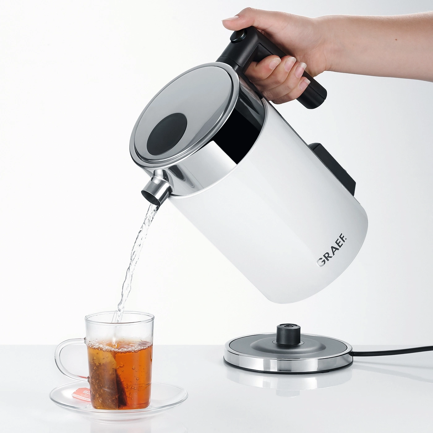 Eine Hand gießt Wasser aus einem weißen Wasserkocher in eine Teetasse.