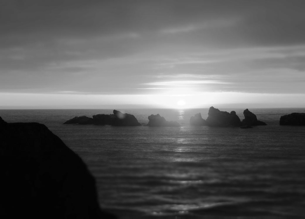 Schwarzweiß-Landschaft mit Meer, Felsen und Sonnenuntergang am Horizont.
