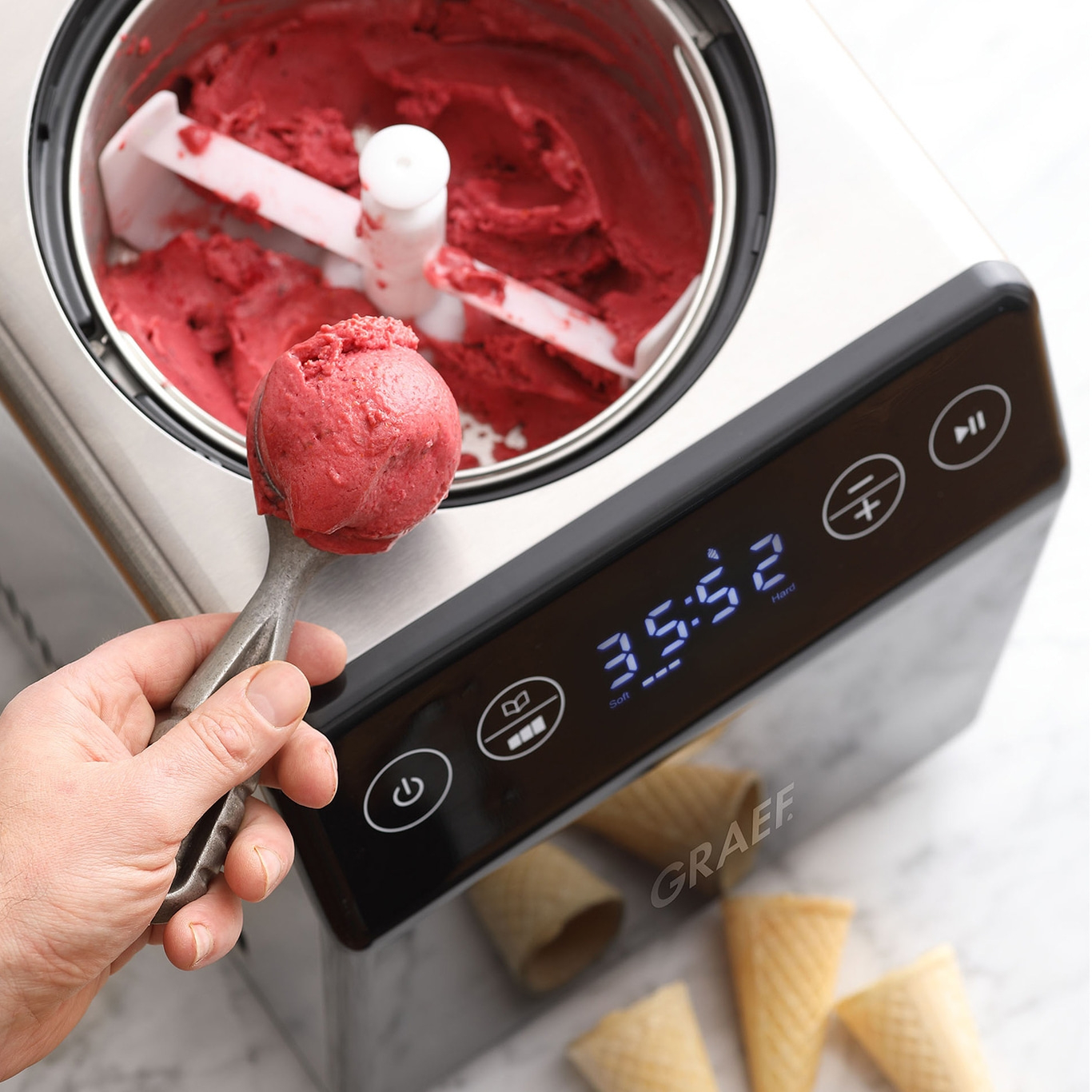 Eiscreme-Maschine mit rotem Eis, Eisportionierer und Waffelhörnchen.