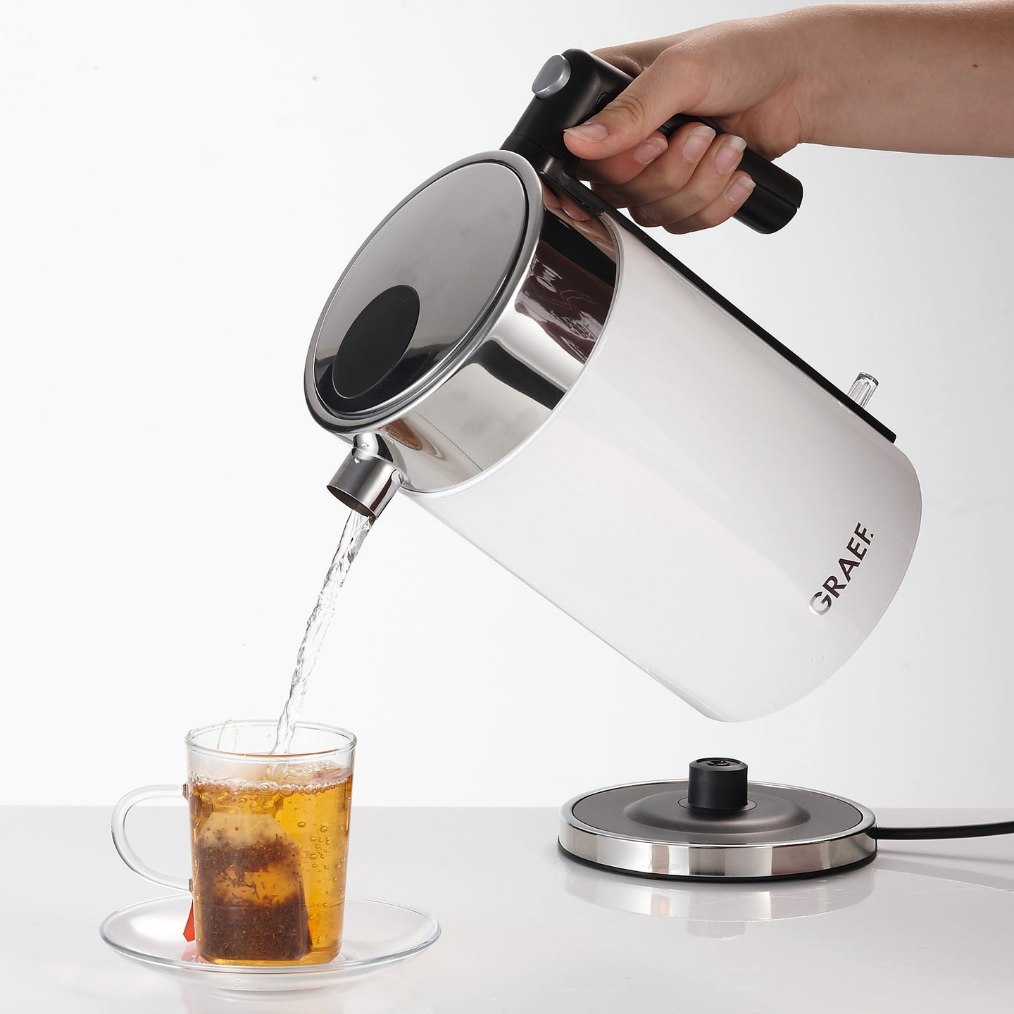 Eine Hand gießt heißes Wasser aus einem Wasserkocher in eine Teetasse mit Teebeutel.