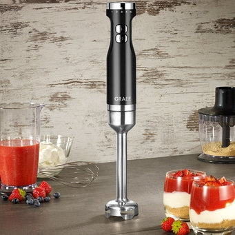 Schwarzer Stabmixer mit Früchten und Desserts auf einem Tisch.