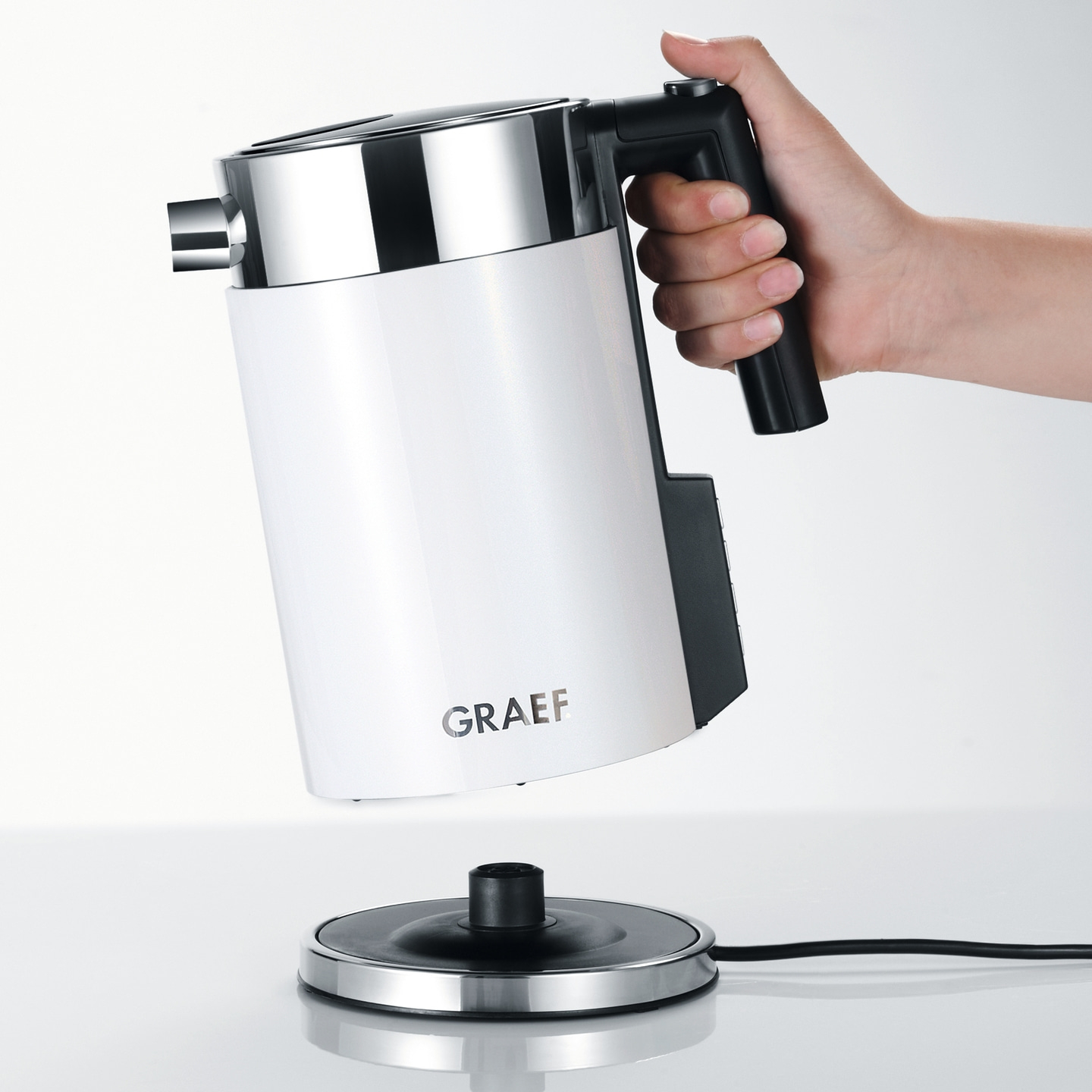 Weißer Wasserkocher mit Griff, der von einer Hand gehalten wird, über einer Basis.
