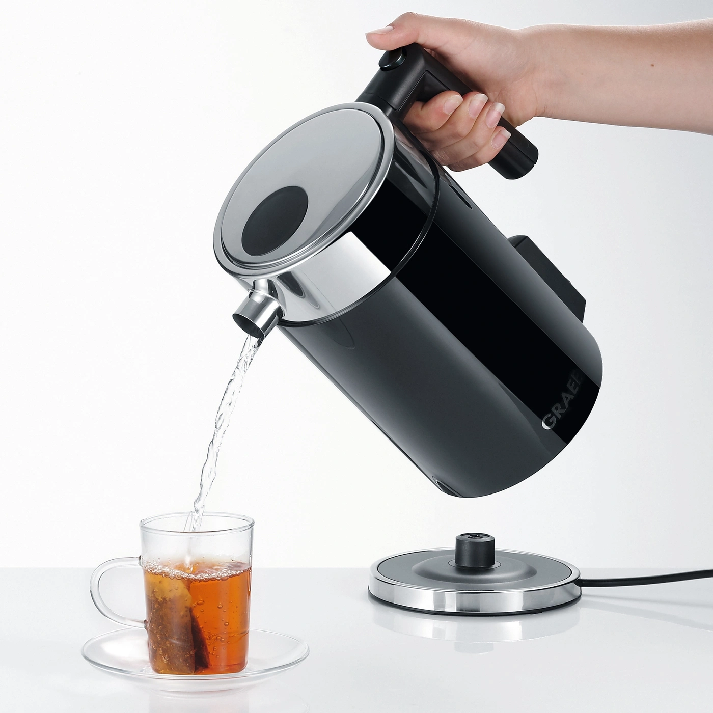 Eine Hand gießt Wasser aus einem schwarzen Wasserkocher in eine Teetasse.