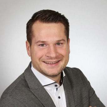 Ein lächelnder Mann in einem grauen Blazer und weißem Hemd.