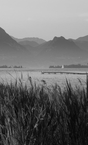 Landschaftsfoto mit Bergen, See, Steg und Segelboot in Schwarzweiß.