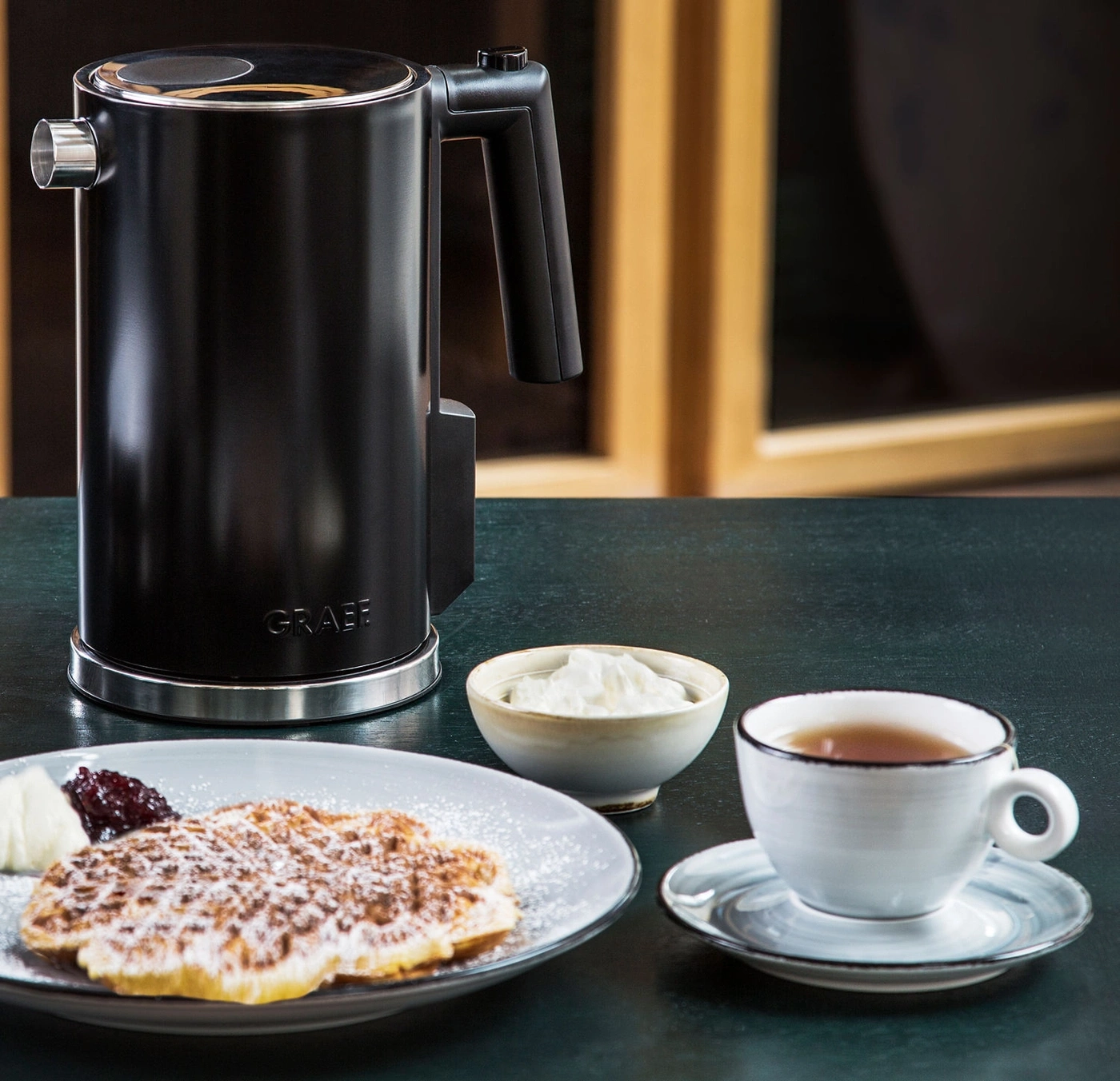 Schwarzer Wasserkocher, Waffel, Sahne, Marmelade und Tee auf dem Tisch.