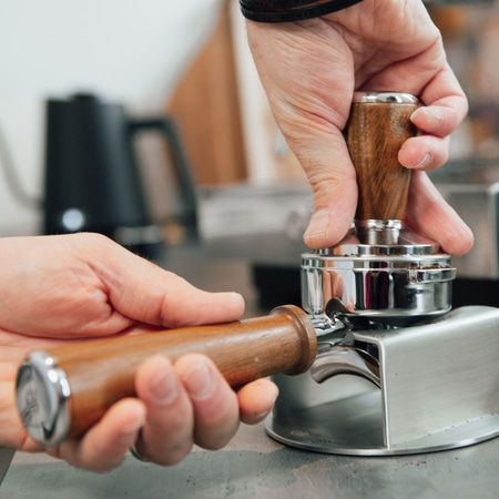 Hände drücken Kaffeepulver mit einem Tamper in einem Siebträger zusammen.