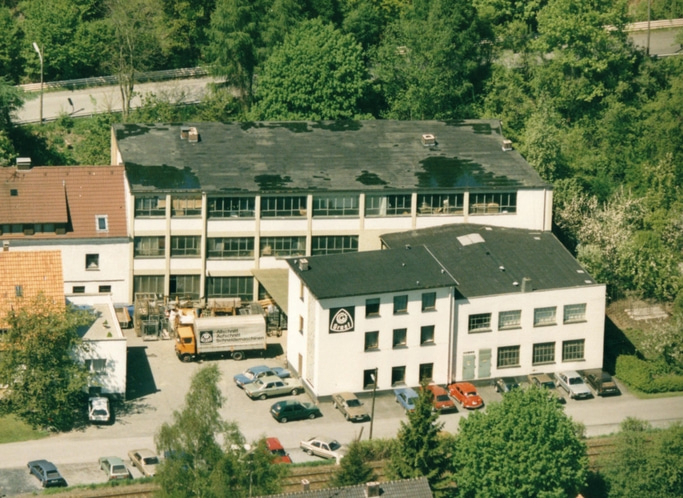 Luftaufnahme eines Fabrikgebäudes mit Parkplätzen und Bäumen in der Nähe.