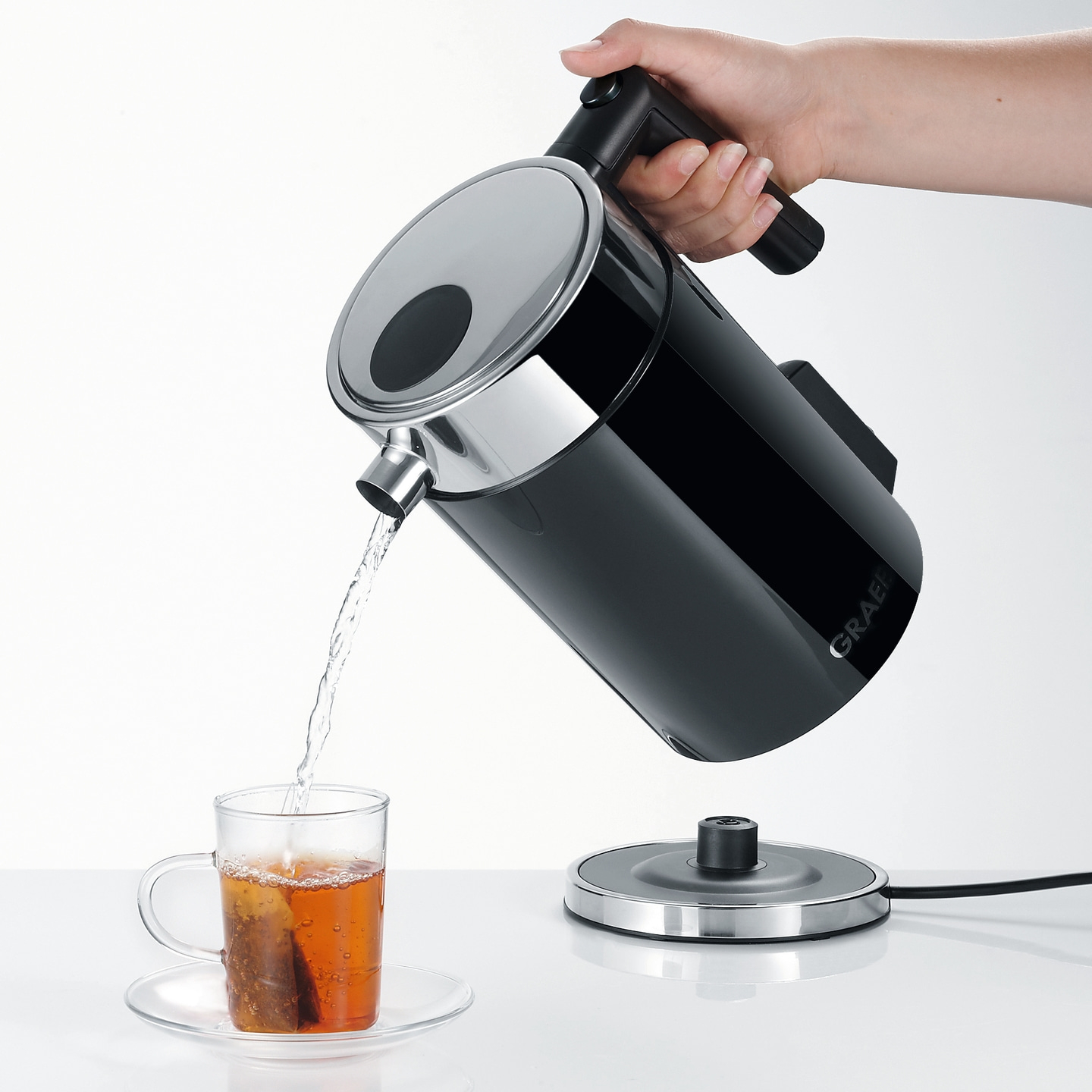 Eine Hand gießt Wasser aus einem schwarzen Wasserkocher in eine Teetasse.
