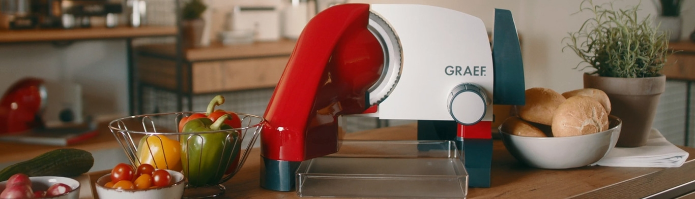 Scène de cuisine avec trancheuse, légumes et pain sur une table en bois.