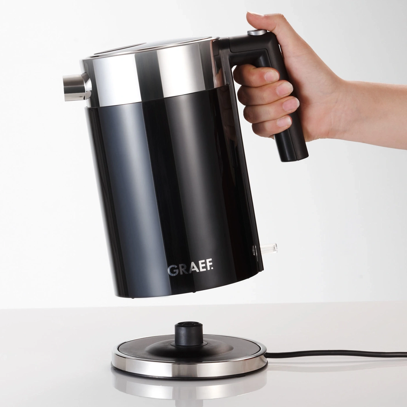 Schwarzer Wasserkocher mit Griff, der von einer Hand gehalten wird, über einer Basis.