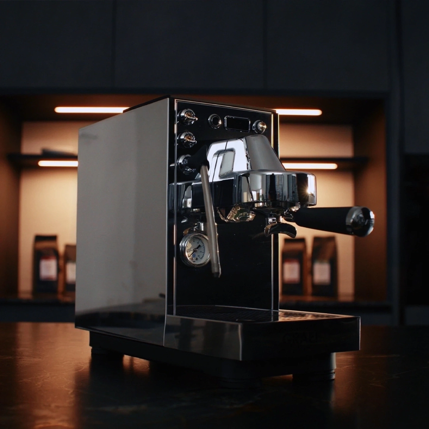 Eine glänzende Espressomaschine steht auf einem dunklen Tisch, im Hintergrund sind Kaffeepackungen.