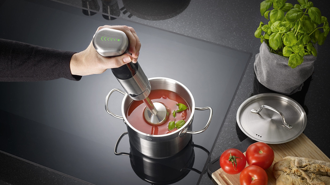 Person püriert Suppe mit einem Stabmixer in einem Topf mit Tomaten und Basilikum.