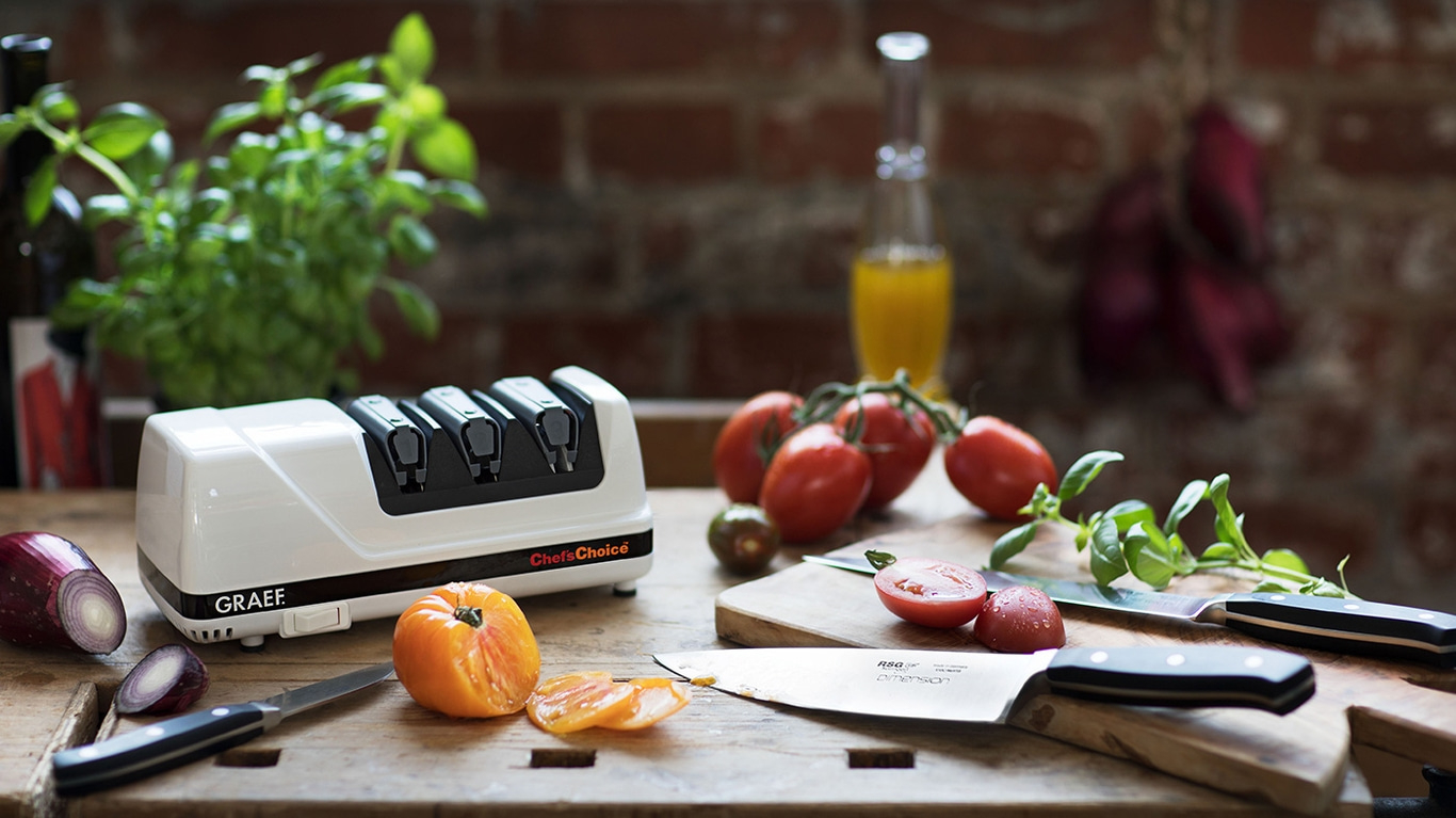 Küchenszene mit Tomaten, Messer, Schleifgerät und Zwiebeln auf einem Holztisch.