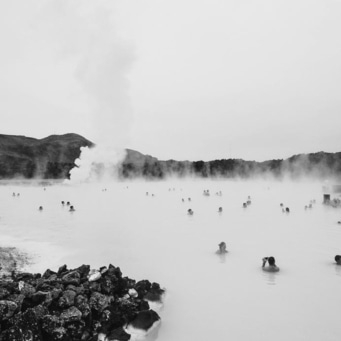 Menschen baden in einem milchig-blauen See mit Dampf und Bergen im Hintergrund.