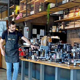 Mann in einer Bar, der auf eine Kaffeemaschine zeigt, mit Regalen im Hintergrund.