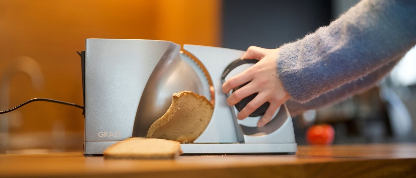 Person schneidet Brot mit einer Brotschneidemaschine auf einer Holzoberfläche.