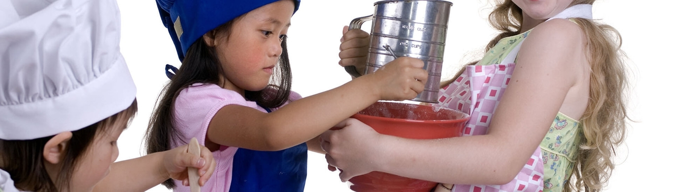 Des enfants qui cuisinent ensemble, une fille tamise de la farine dans un bol.