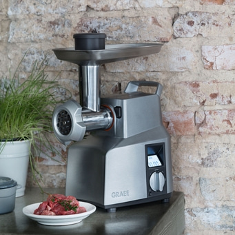 Meat grinder with meat on a plate in front of a brick wall background.