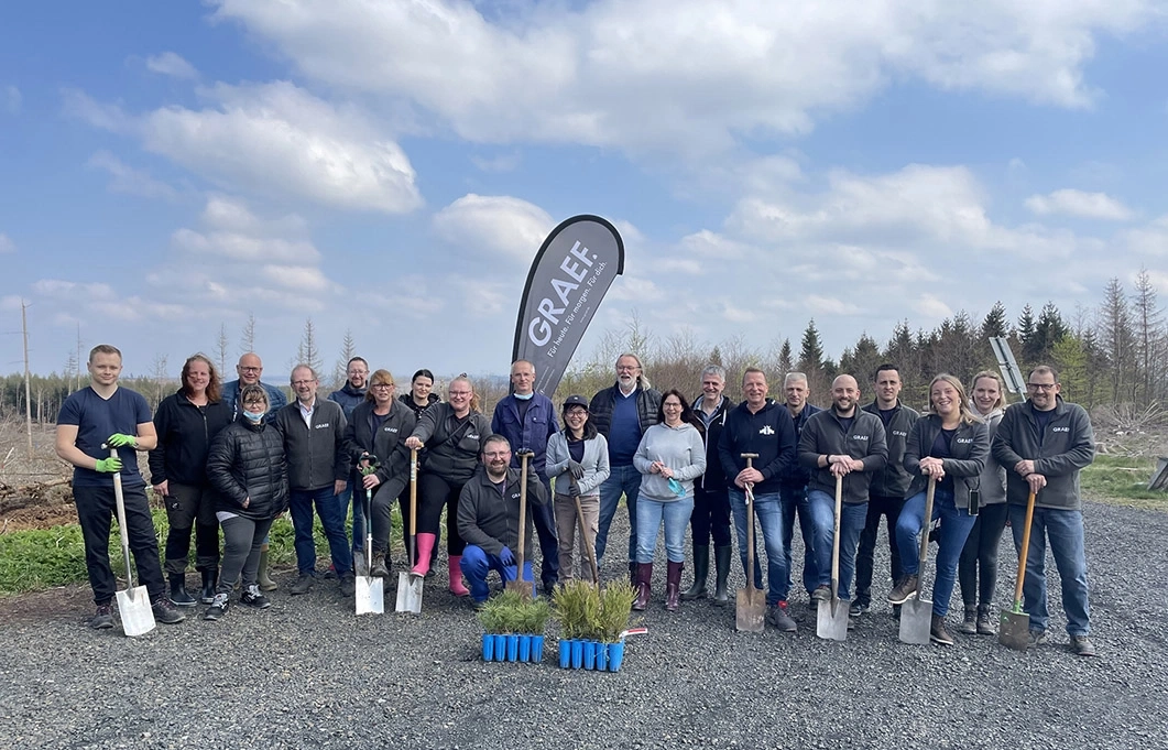 Gruppe von Menschen mit Spaten vor Bäumen und blauem Himmel.