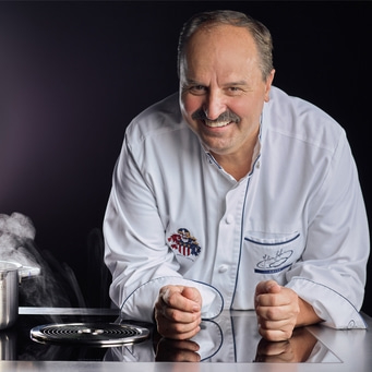 Ein Koch in weißer Jacke, der sich auf eine Kochplatte stützt und lächelt.