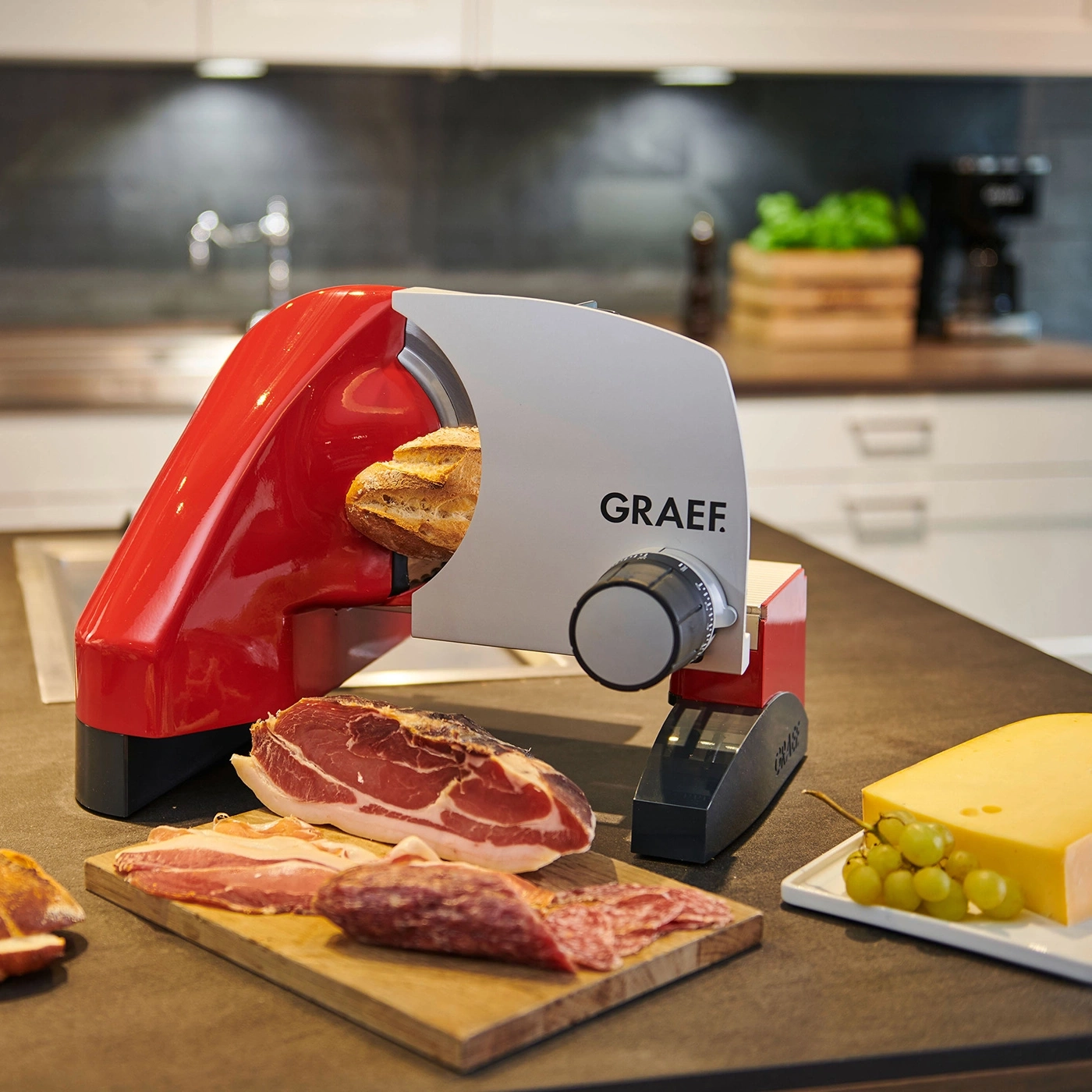 Roter Aufschnittschneider mit Brot, Wurst, Käse und Trauben auf einer Küchenarbeitsplatte.
